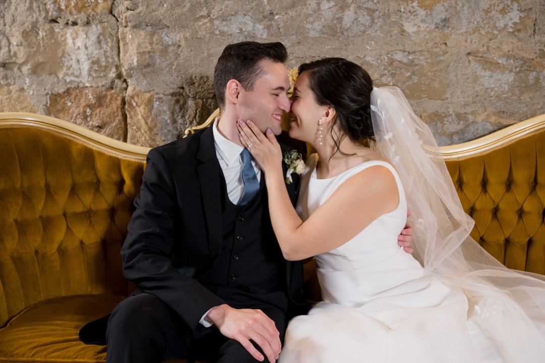 A bride and groom sit on a vintage yellow couch in front of a stone wall at The Art Room Wedding. The bride, in a white gown and veil, gently holds the grooms face as they smile at each other, exuding joy and affection. The groom is dressed in a dark suit and tie.