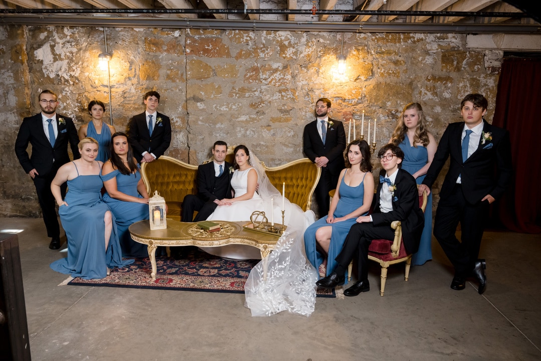 The bride and groom sit on a vintage sofa in The Art Room Wedding venue, surrounded by their party. Bridesmaids wear light blue dresses, and groomsmen sport black suits with blue ties. Theyre enveloped by rustic charm with exposed stone walls and warm lighting.