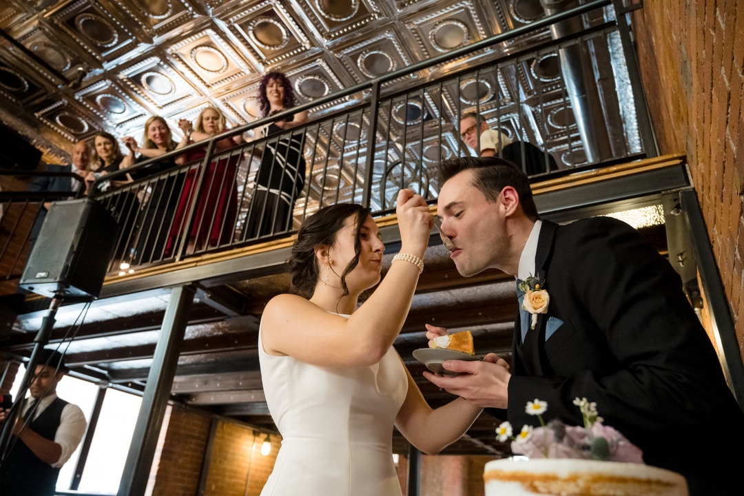 At The Art Room Wedding, the bride and groom share a slice of cake, with the bride feeding the groom. They stand in a warmly lit venue with exposed brick walls and a decorative ceiling. Guests watch from a balcony above, smiling and capturing the moment.