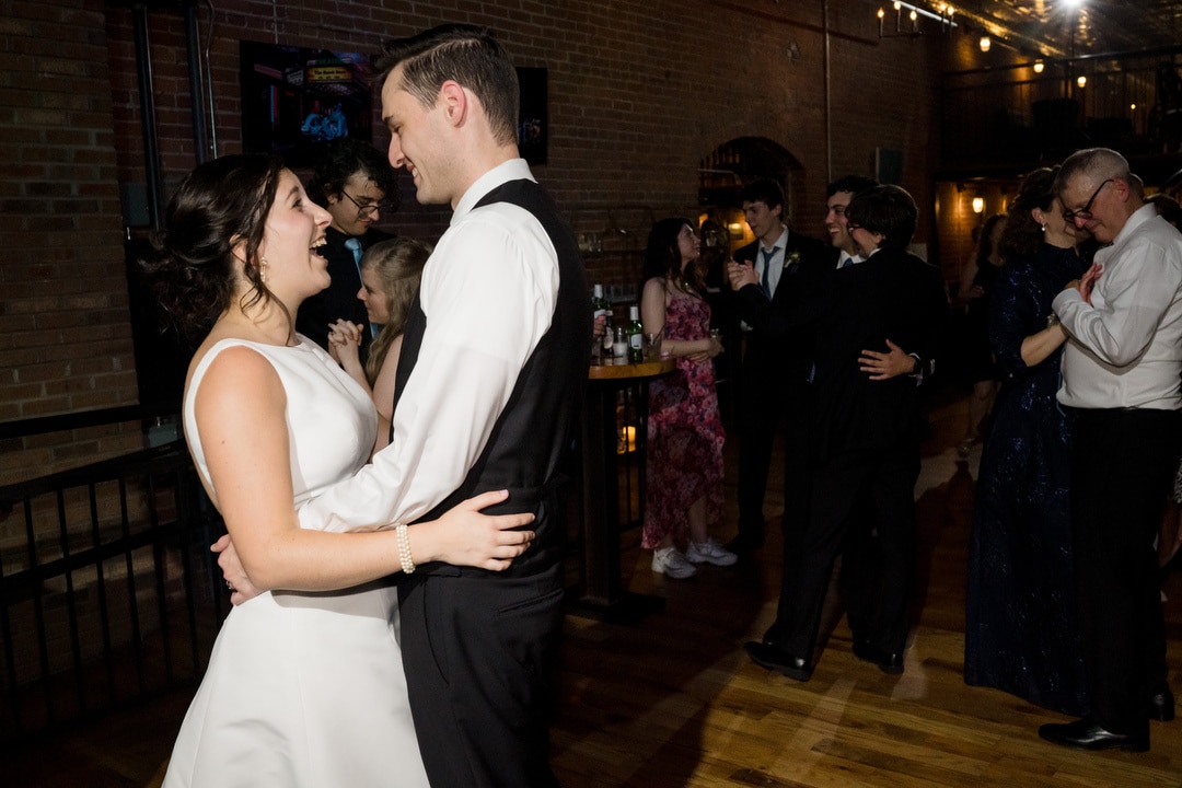 In the dimly lit elegance of The Art Room Wedding, a couple in formal attire dances closely on wooden floors, smiling at each other. Around them, other attendees in various outfits sway and mingle beneath twinkling string lights hanging from the ceiling.