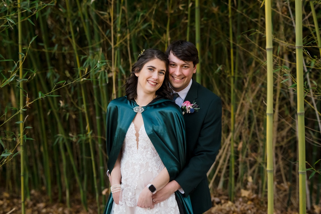 In a serene bamboo forest, a couple stands close; the woman in a white dress and dark green cape, the man in a matching suit adorned with a boutonniere. Their smiles radiate joy reminiscent of their Pittsburgh wedding at Saint-Anne-Church, as they embrace amid towering bamboo stalks.
