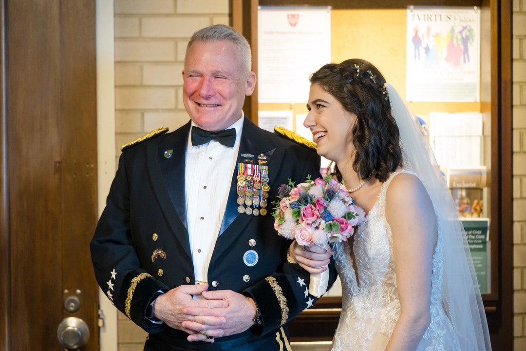 A man in a decorated military uniform smiles alongside a joyful bride with a bouquet, standing indoors by a bulletin board. The bride, in her white dress and veil, might be reminiscing about their beautiful Pittsburgh wedding at Saint-Anne-Church.