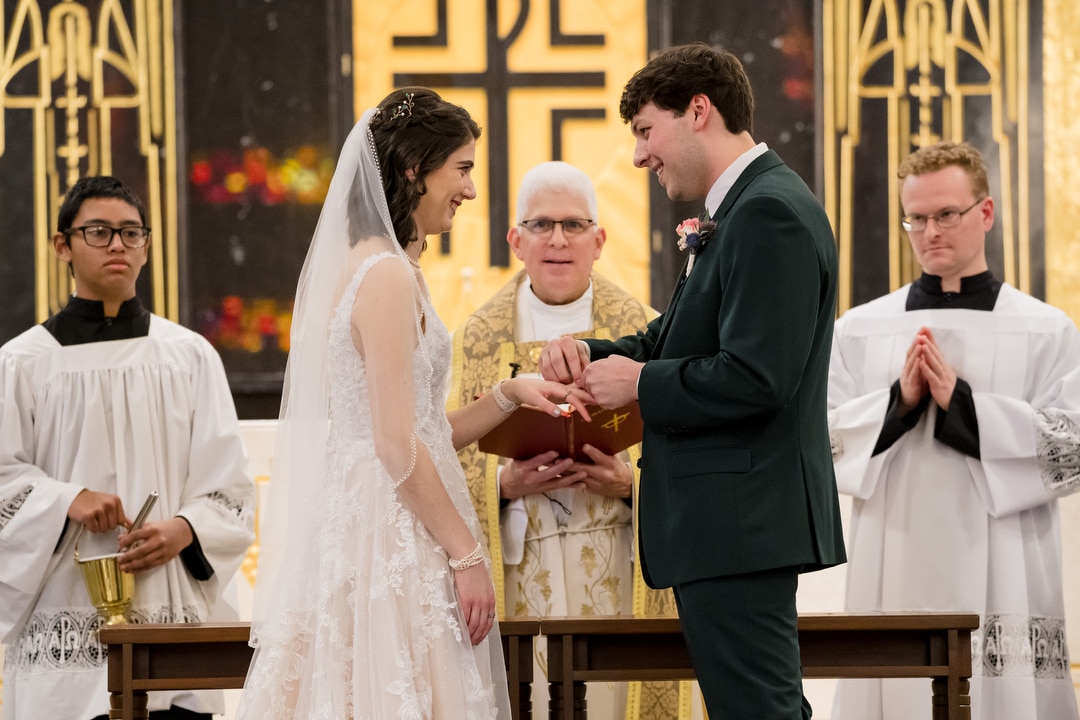 At Saint-Anne-Church in Pittsburgh, a bride and groom lovingly exchange rings during their wedding ceremony, smiles lighting up their faces. The priest stands between them with a book, flanked by two altar servers, all set against the backdrop of elaborate church decor.