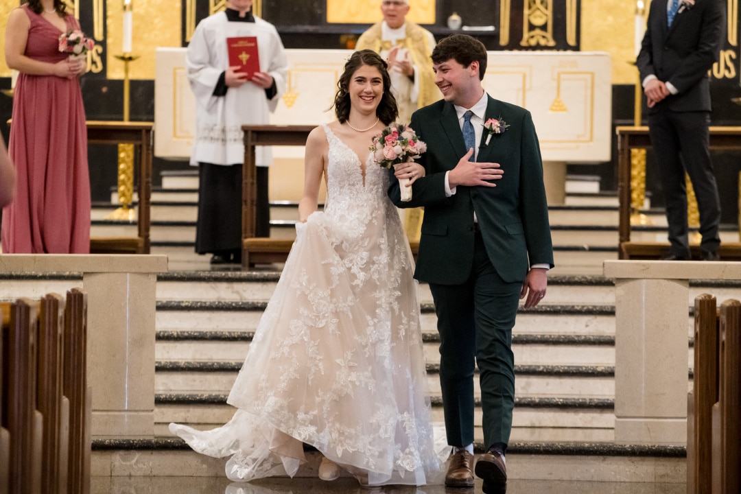 The bride and groom smile joyfully as they walk down the aisle of Saint-Anne-Church in Pittsburgh. The bride, in a lace gown holding a bouquet, and the groom, in a dark suit, are surrounded by the beauty of an ornate altar.