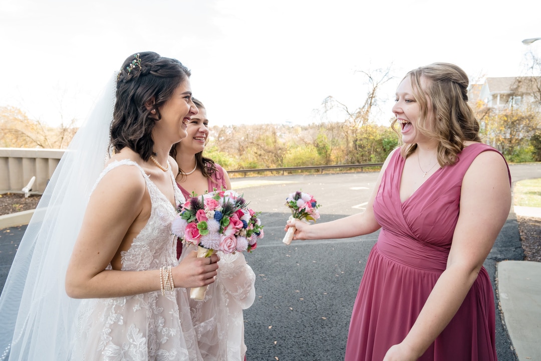 The bride and her two bridesmaids in pink dresses are outdoors, beaming as they hold their bouquets. With a floral veil draped elegantly, they stand on a paved area with bare trees and a railing behind them near Saint-Anne-Church in Pittsburgh.