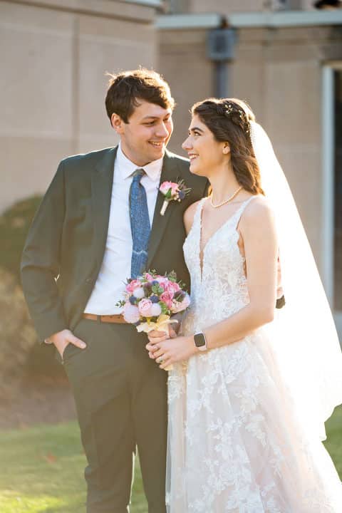 A bride and groom stand outdoors in Springwood on a sunny day. The bride wears a lace wedding dress and veil, holding a bouquet of pink and purple flowers. The groom, with a pink boutonniere, is dressed in a dark suit. Theyre gazing at each other and smiling joyfully in this picturesque Pittsburgh setting.