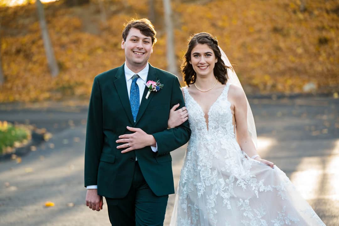 A bride in a white lace gown and veil and a groom in a dark suit with a pink boutonniere are happily walking arm in arm through Springwood. The blurred autumn scene, reminiscent of Pittsburghs charm, features leaves scattered gracefully on the ground.