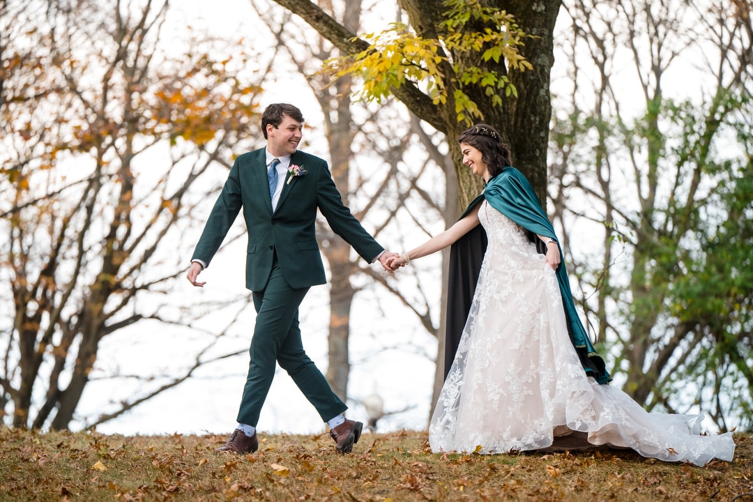 A couple holds hands and walks outdoors on an autumn day in Springwood. The person in a white lace dress wears a green shawl, while the one in a dark suit smiles at them. Fallen leaves and trees with sparse foliage surround their peaceful retreat near Pittsburgh.