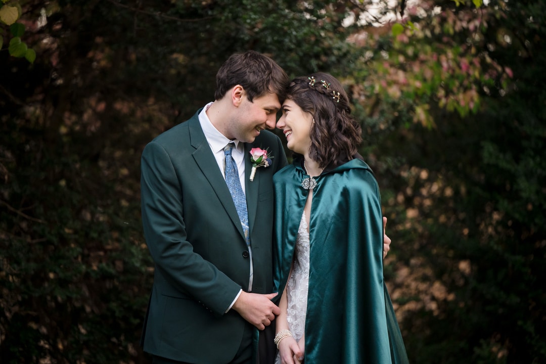 In the charming setting of Springwood, a couple stands close, smiling and touching foreheads. The man wears a green suit with a boutonniere, while the woman dons a green cape adorned with floral details. The backdrop of trees adds to the romantic ambiance of this Pittsburgh wedding scene.