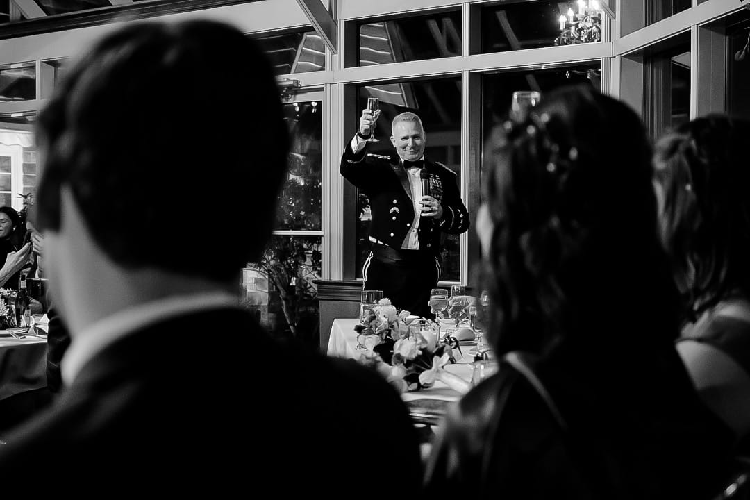 A man in a formal military uniform raises a glass for a toast at the Springwood wedding dinner. He stands near a long table adorned with floral arrangements. Blurred guests in the foreground listen attentively. The setting, with large windows, evokes elegance. Black and white.