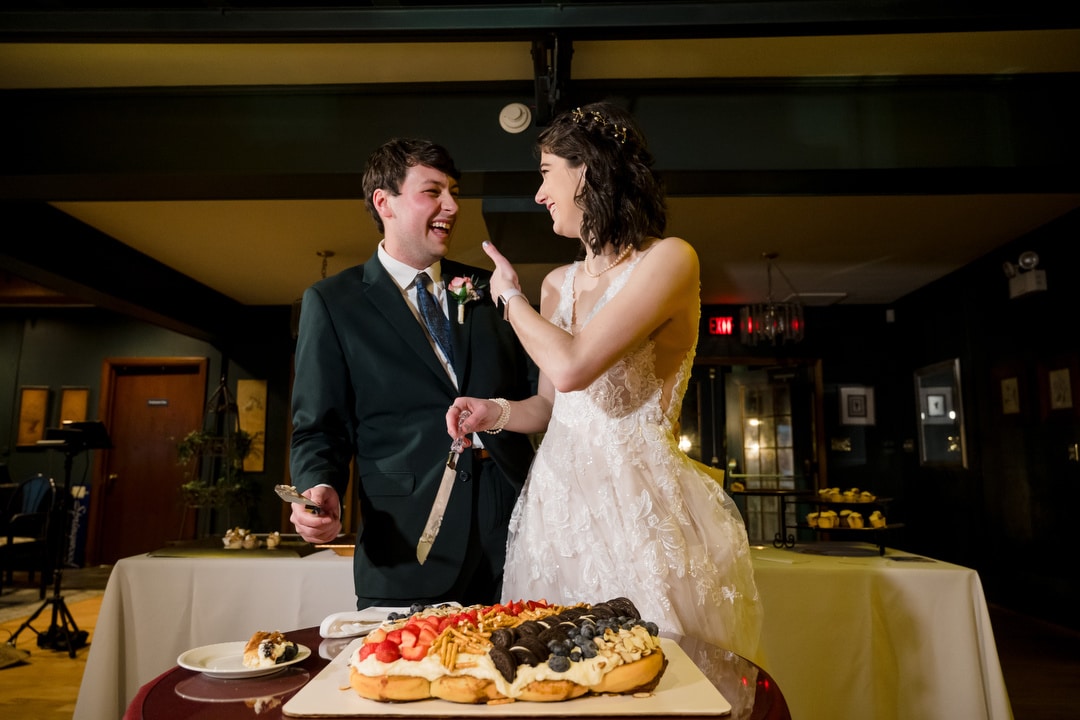 In a warmly lit room in Springwood, a bride playfully feeds her groom with a fork. They stand before a table adorned with a vibrant dessert platter. The groom holds his plate, and both share joyful smiles, capturing the essence of their Pittsburgh wedding celebration.