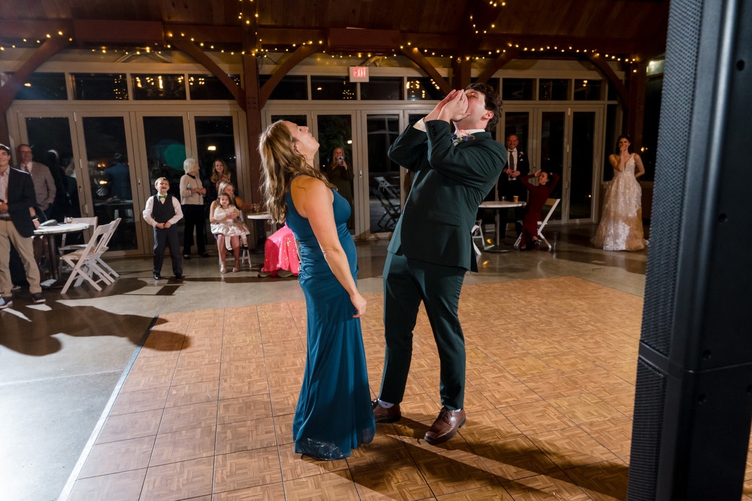 A woman in a blue dress and a man in a suit are on the dance floor at a Springwood wedding. The man shouts playfully while the woman laughs under the twinkling string lights. In Pittsburgh, their joy is contagious as people in the background watch and smile.