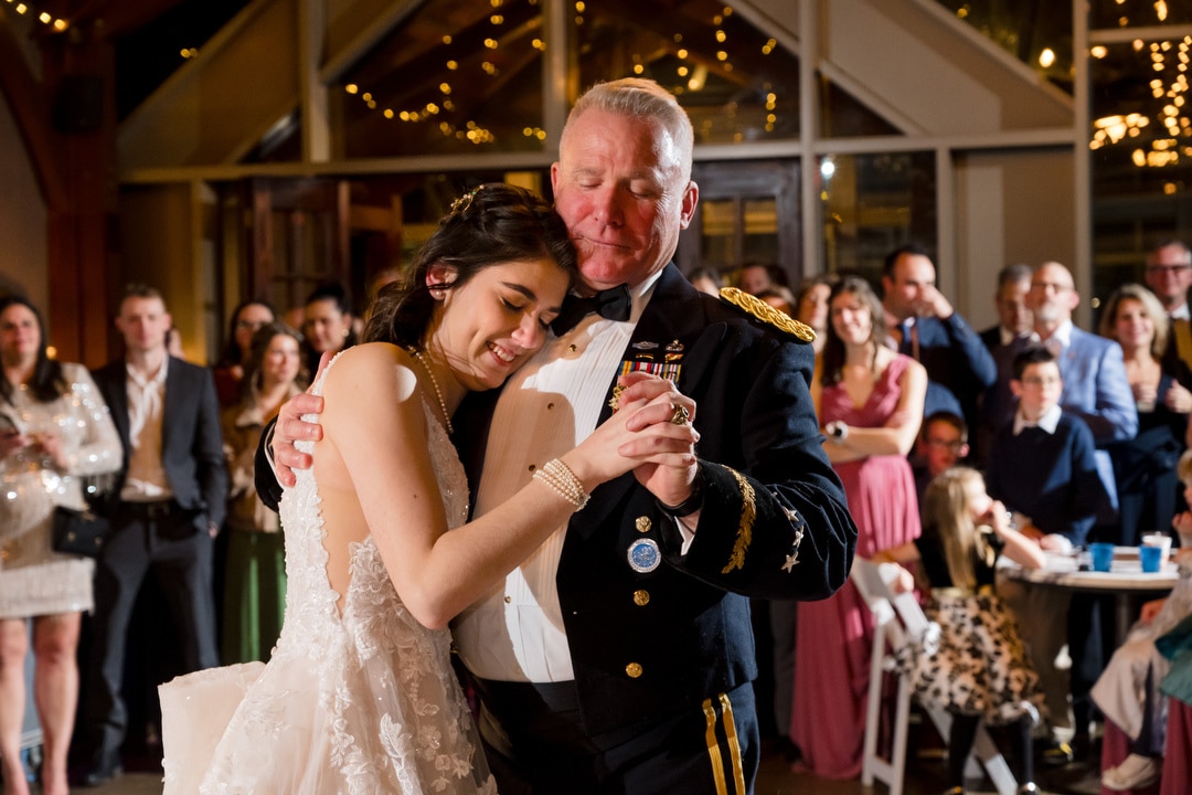 At a wedding reception in Springwood, a bride in a white dress joyfully dances with a man in a decorated military uniform. Surrounded by smiling guests beneath warm string lights, the Pittsburgh evening feels magical and intimate.