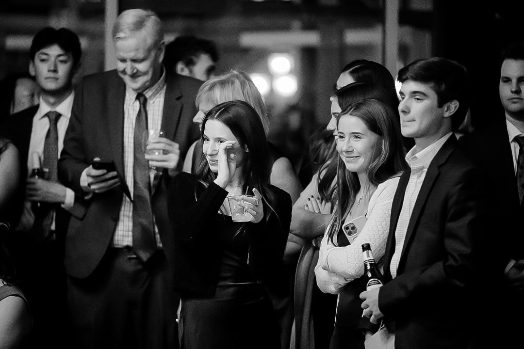 A group of people in formal attire gather at an indoor event in Pittsburgh. They are smiling, some holding drinks, while one man glances at his phone. The black-and-white image captures the essence of a social gathering that could easily be a wedding celebration.