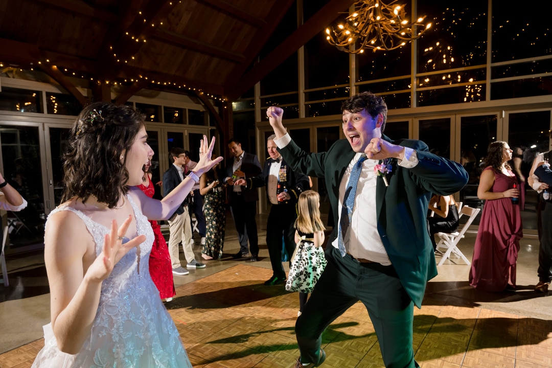 A bride in a white dress and a groom in a suit dance energetically on the wooden floor at their Springwood wedding reception. Guests in formal attire watch and join the dance under string lights and a chandelier, making it an unforgettable evening near Pittsburgh.