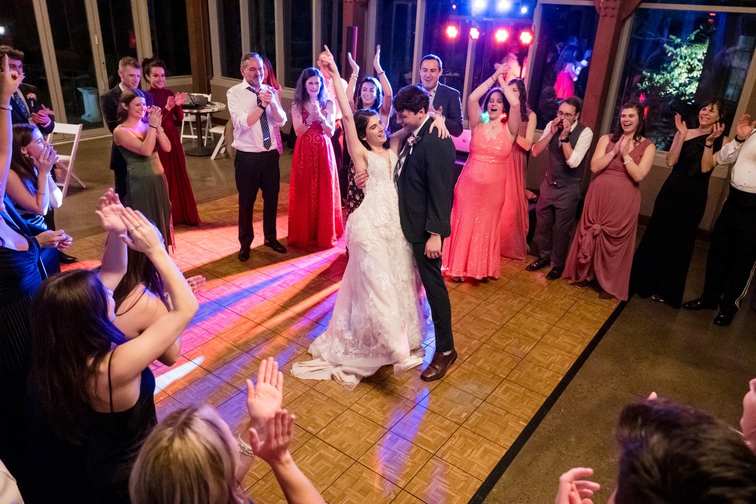 At a charming Springwood wedding, a couple shares a joyful dance surrounded by guests on the wooden floor. The bride wears a white gown, and the groom a dark suit. Attendees in formal attire cheer as colorful lights illuminate this lively Pittsburgh scene.