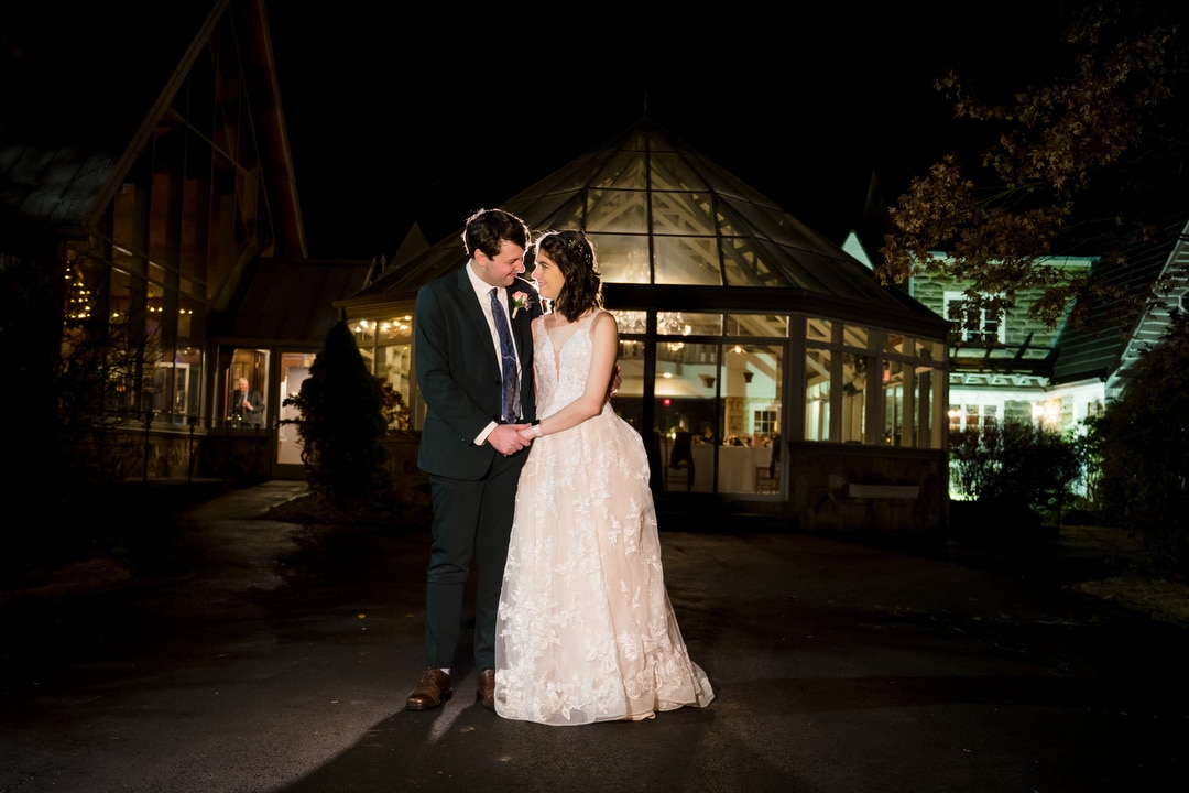 A couple in wedding attire stands hand in hand on a dimly lit path. The woman is in a white gown and the man in a dark suit. They stand before Springwoods brightly lit pavilion, with trees nearby. Pittsburghs night sky provides a stunning backdrop for this idyllic moment.
