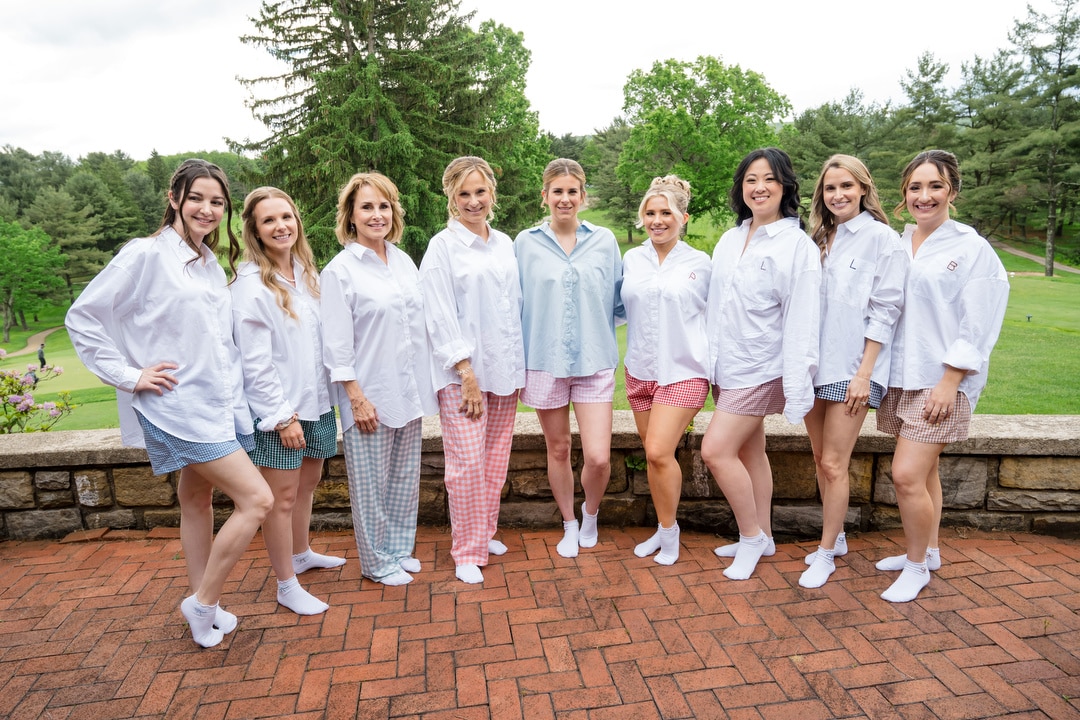Nine women wearing oversized button-up shirts and shorts stand side by side on a brick patio outdoors, smiling at the camera with green trees and grass in the background.