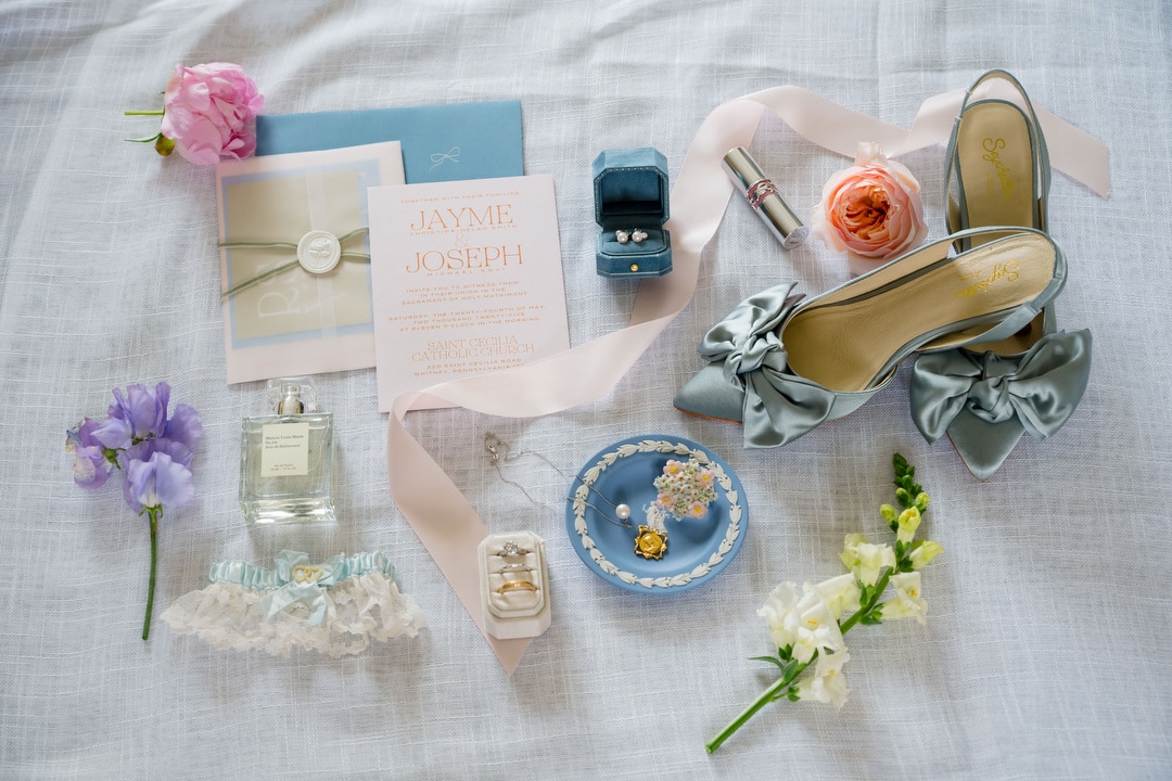 Flat lay of wedding items including invitations, blue high heels with bows, jewelry, flowers, garter, perfume, lipstick, and a ring box, all arranged on a light fabric background.