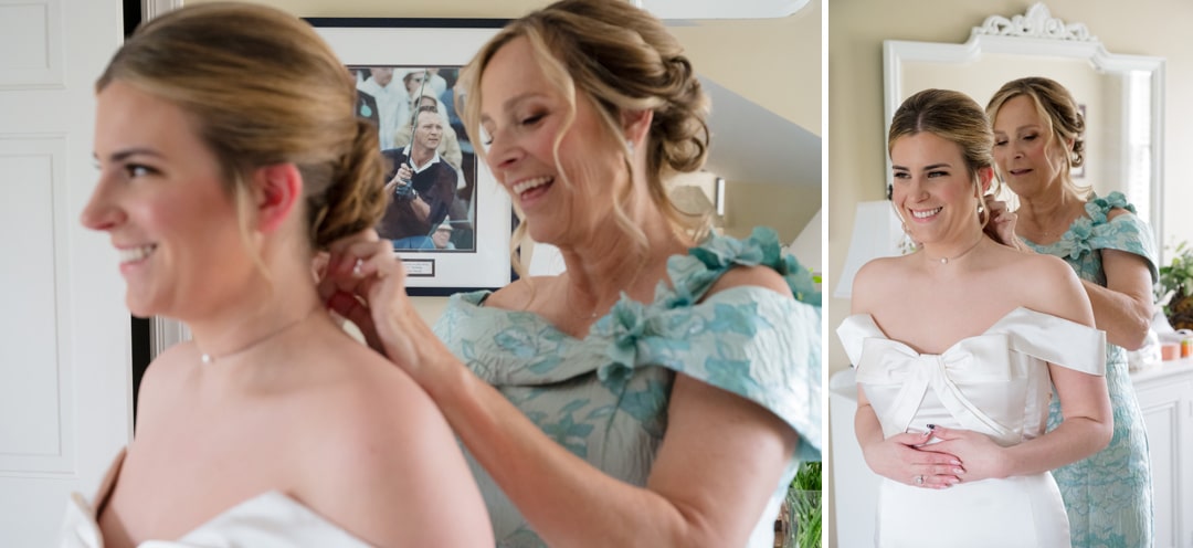 A woman in a light blue dress helps a smiling bride adjust her necklace. They stand in a bright room with a mirror and framed photo on the wall. The bride wears an off-shoulder white gown. Both look happy and excited.
