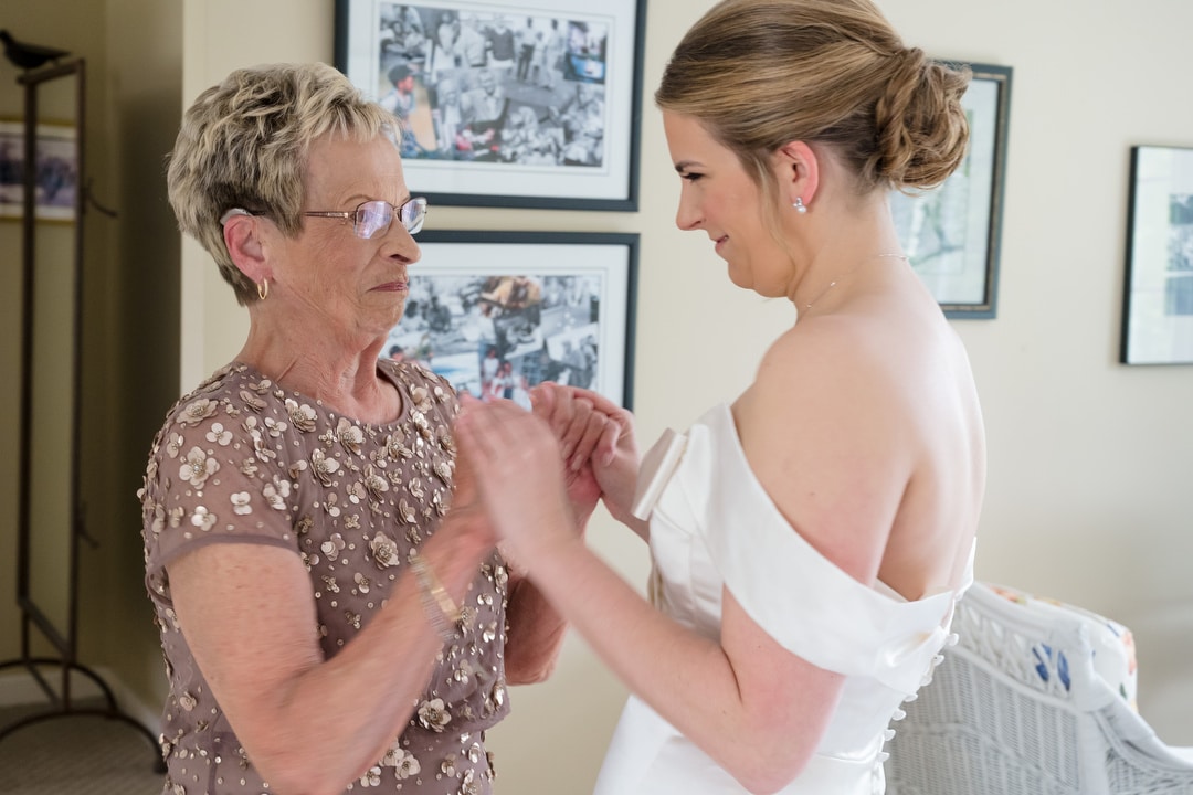 An older woman and a bride in a white dress smile and hold hands indoors, sharing a tender moment before the wedding. Framed photos hang on the wall behind them.