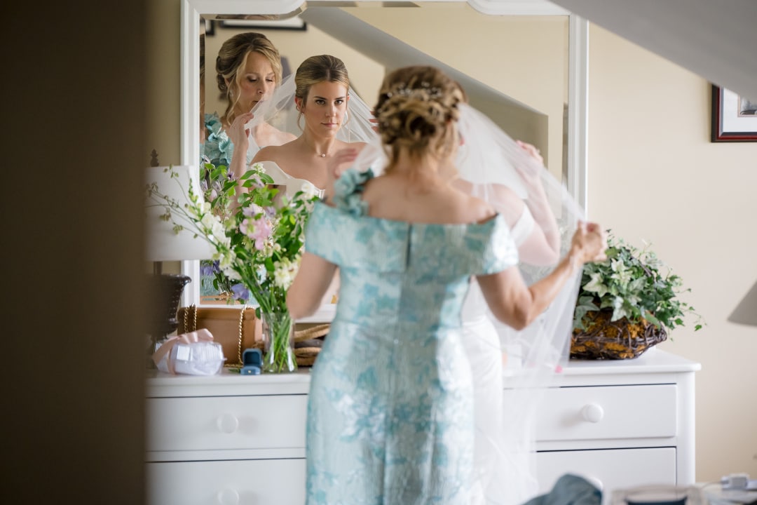 A bride stands in front of a mirror as another woman helps adjust her veil. The brides reflection shows her focused expression. Flowers and decorative items sit on the white dresser below the mirror.