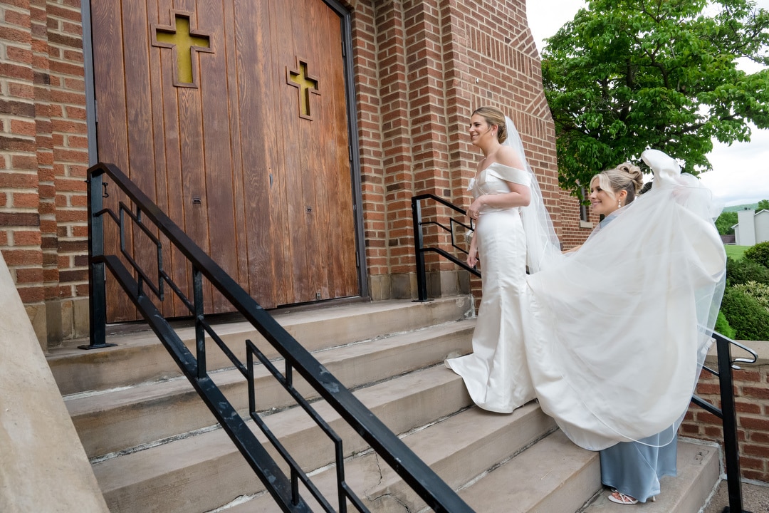 A bride in a white dress walks up church steps as a bridesmaid holds her train. The wooden church doors have cross-shaped windows, and there is greenery in the background.