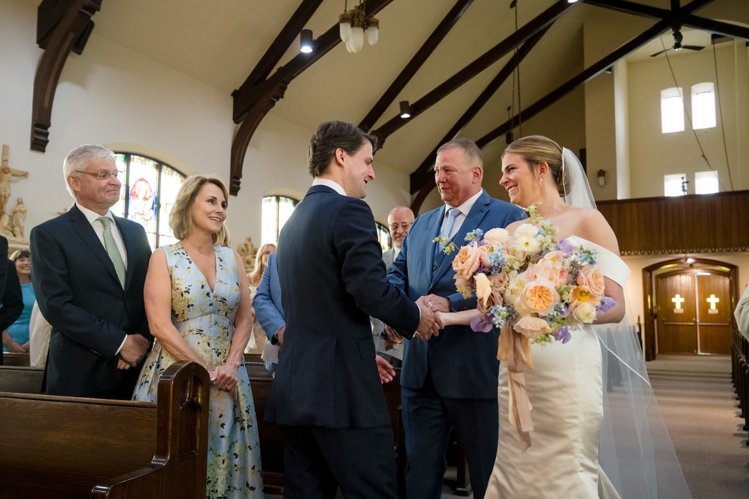 A bride in a white dress smiles as she holds a bouquet and shakes hands with a man in a suit inside a church. A man and woman, likely her parents, stand nearby, smiling. Guests are seated in the background.