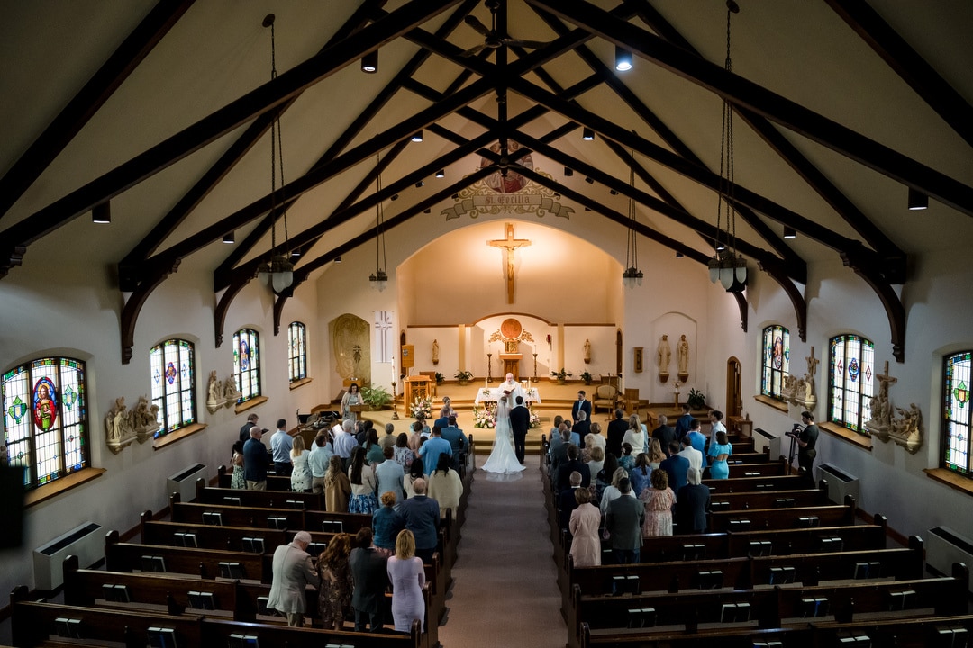 A bride and groom stand at the altar in a church filled with seated guests. The vaulted ceiling, exposed beams, stained glass windows, and religious statues create a serene wedding ceremony setting.