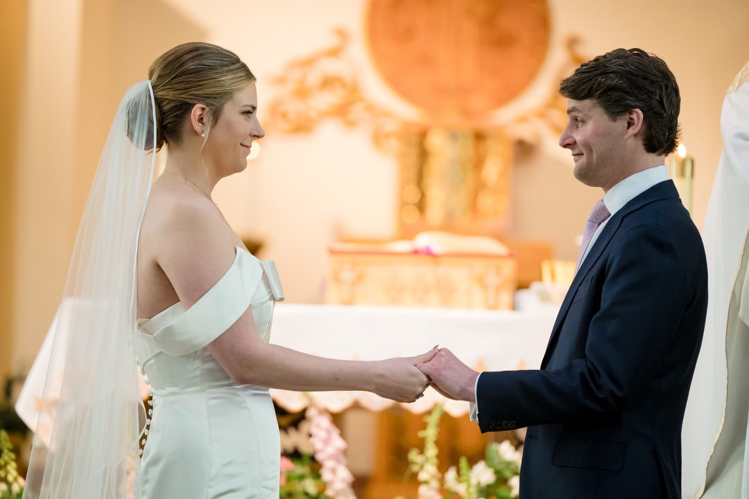 A bride and groom stand facing each other, holding hands and smiling, during a wedding ceremony in a church decorated with flowers and a golden altar in the background.