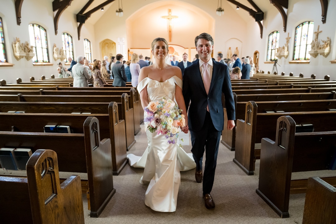 A bride in a white gown holding a colorful bouquet and a groom in a suit walk smiling down the aisle of a church with guests standing and wooden pews on either side.