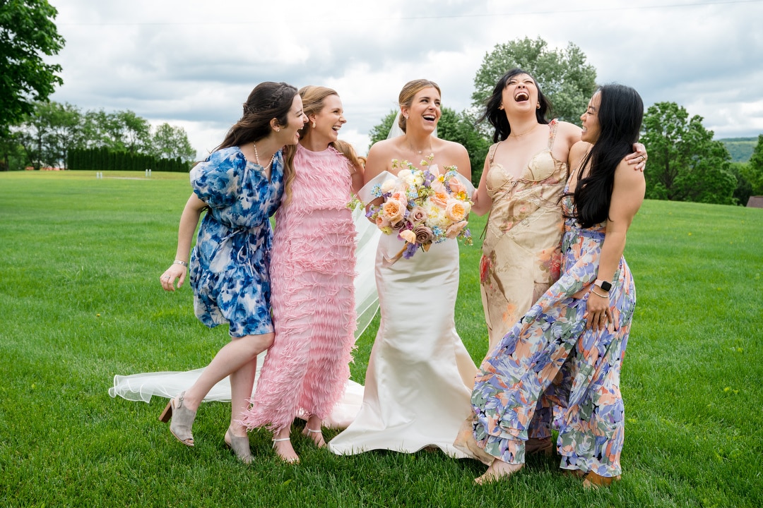 Five women, including a bride in a white gown holding a bouquet, stand arm-in-arm on grass, laughing and smiling, with green trees and a cloudy sky in the background. The others wear colorful dresses.