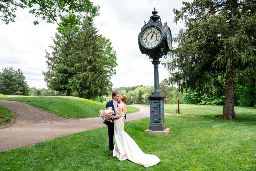 A bride and groom embrace on a green lawn next to a large vintage clock, surrounded by trees and a winding path, on a bright, cloudy day.