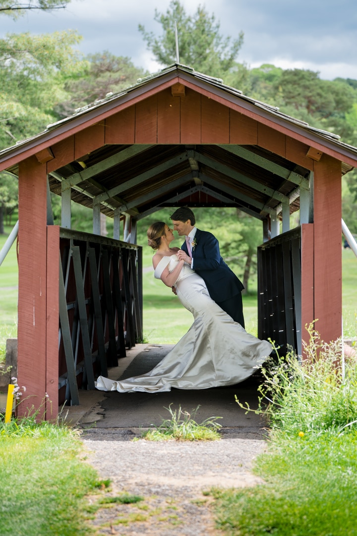 A bride and groom in wedding attire share a romantic dip and gaze into each others eyes on a red covered wooden bridge surrounded by green grass and trees.