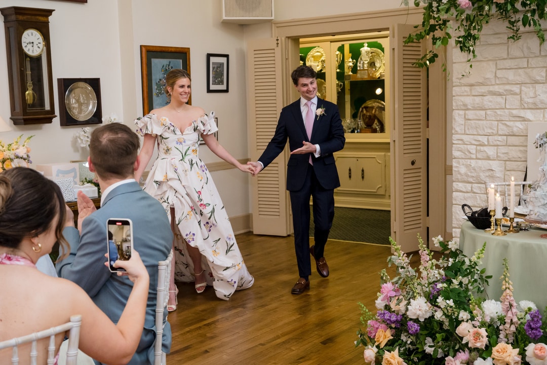 A smiling couple, dressed formally, enters a decorated room holding hands as guests take photos. The room features flowers, candles, and vintage decor, creating a festive atmosphere.