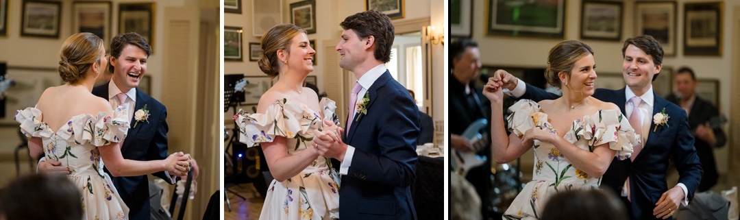A bride and groom dance together, smiling and laughing, in a warmly lit room decorated with framed photos. The bride wears a floral off-the-shoulder dress; the groom wears a navy suit and pink tie.