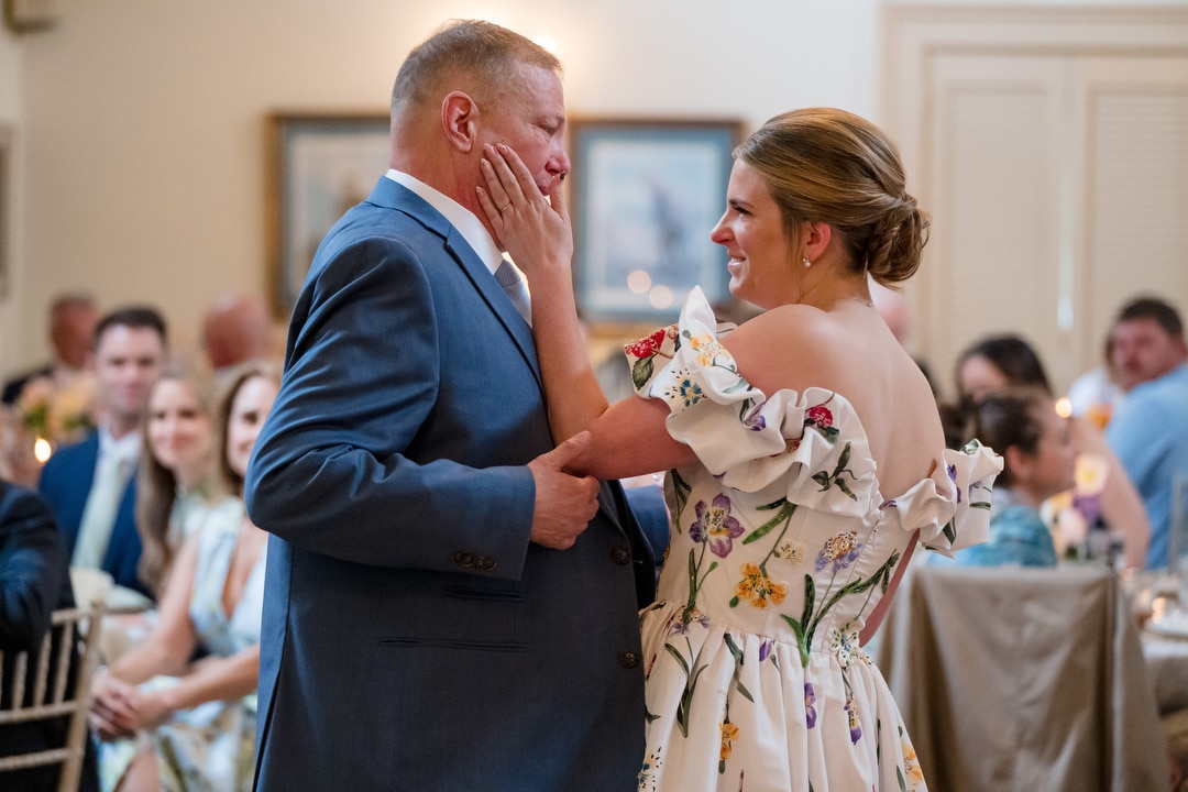 A woman in a floral dress gently touches the face of a man in a blue suit as they dance together at a formal event, surrounded by seated guests who are watching them and smiling.