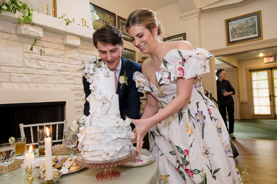 A smiling couple in formal attire cuts a white, floral-decorated wedding cake together at a decorated indoor venue. The woman wears a floral dress, and the man is in a dark suit with a boutonniere.