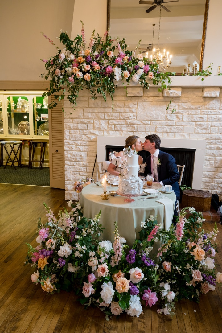 A bride and groom sit at a round table adorned with flowers and a cake, sharing a kiss in front of a decorated fireplace with a large floral arrangement above. The scene is elegant and romantic.
