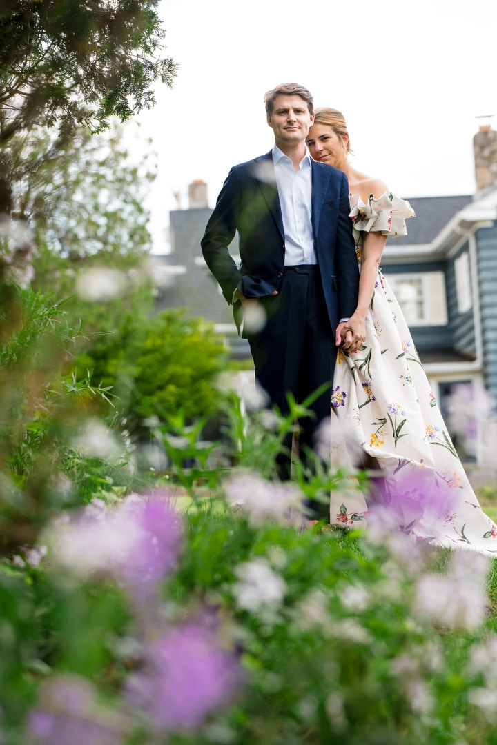 A couple stands together in a garden, holding hands. The woman wears a white floral dress and the man wears a navy suit. Purple flowers and greenery are in the foreground, with a house visible in the background.