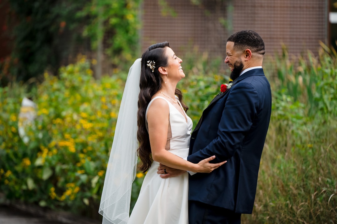 A bride in a white dress and veil and a groom in a navy suit stand outdoors, facing each other and laughing, surrounded by greenery and yellow flowers.