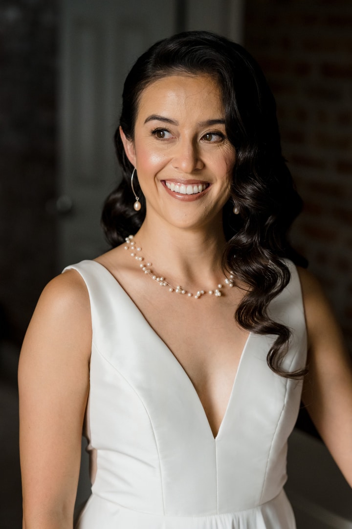 A woman with long, wavy dark hair wearing a white sleeveless dress, pearl necklace, and drop earrings smiles and looks to the side in a softly lit indoor setting.