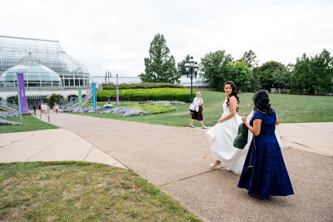 A bride in a white gown walks with a woman in a blue dress on a garden path near a glass conservatory, while other people stroll in the background under a cloudy sky.
