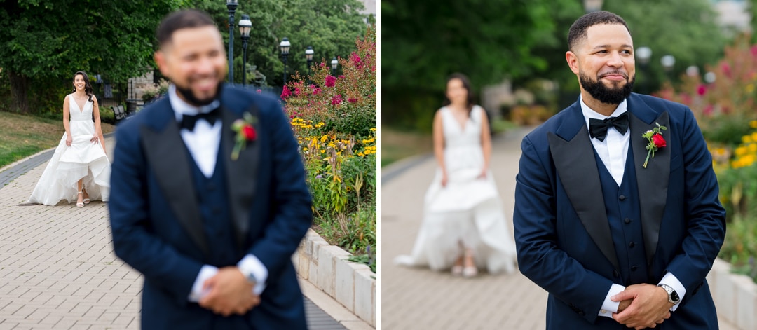 A bride in a white dress approaches a smiling groom in a navy suit and bowtie during an outdoor first look, surrounded by greenery and colorful flowers. The groom stands in focus, while the bride is slightly blurred in the background.