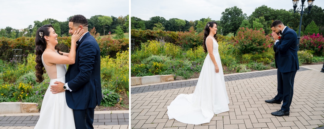 A bride and groom share an emotional first look outdoors; she smiles and holds his face as he reacts with happy tears in a garden setting with greenery and flowers in the background.