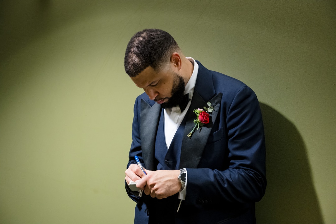 A man in a dark suit with a red rose boutonniere writes on a piece of paper while standing against a plain green wall, looking down and focused on his task.