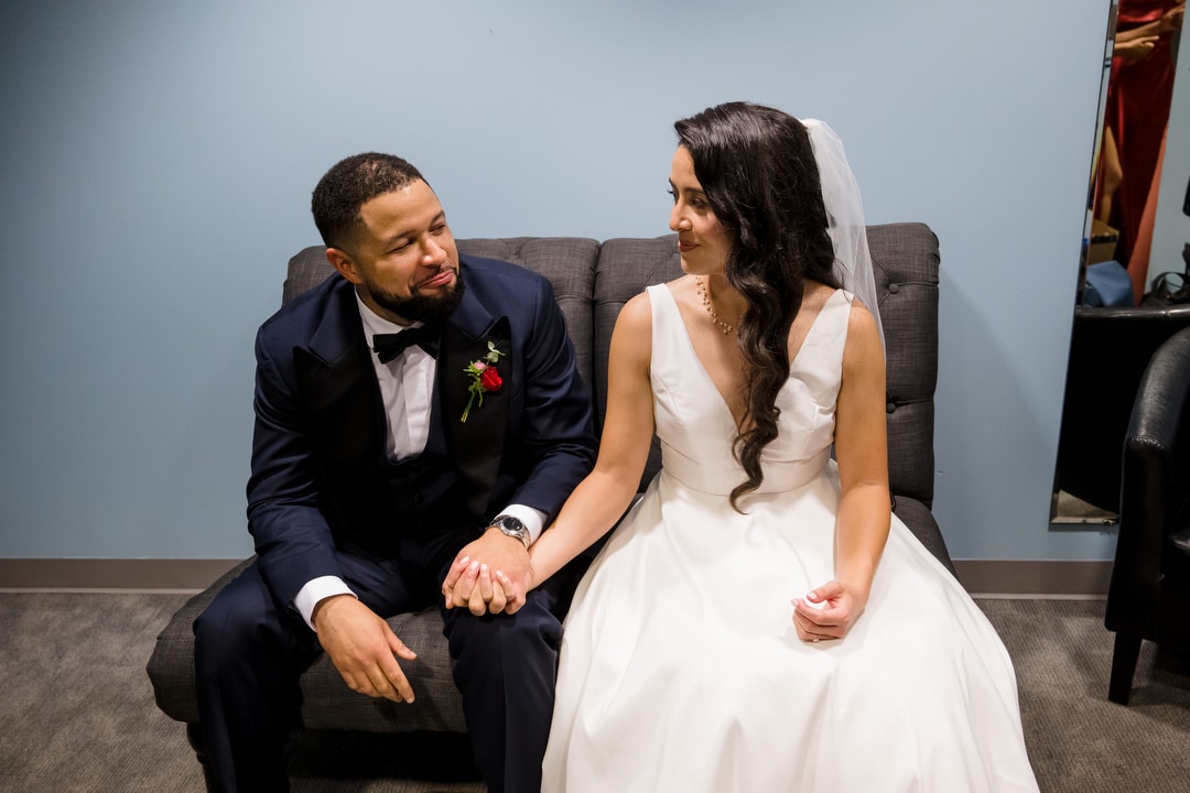 A bride in a white dress and veil holds hands with a groom in a dark suit and bow tie as they sit together on a gray couch, looking at each other and smiling in a softly lit room.