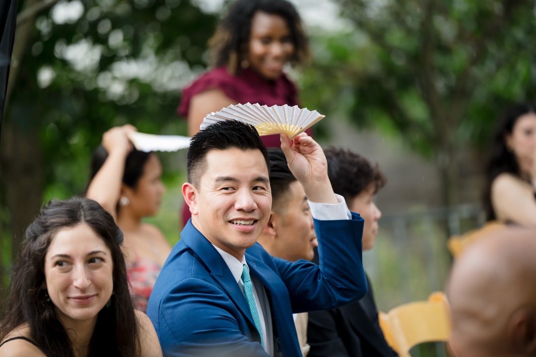 A man in a blue suit smiles at the camera while holding a white fan above his head. Other people around him, seated outdoors, are also using hand fans. Trees and greenery are visible in the background.