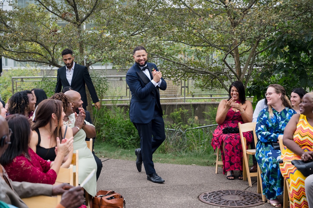 A smiling man in a dark suit walks down an outdoor aisle, clapping his hands, while seated guests on both sides smile, clap, and watch. Another man follows behind. Trees and greenery form the background.