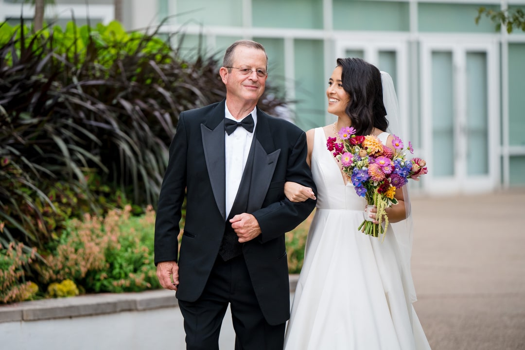 A bride in a white dress holding colorful flowers walks arm-in-arm with an older man in a black tuxedo outdoors, both smiling and looking at each other. Green plants and a modern building are visible in the background.
